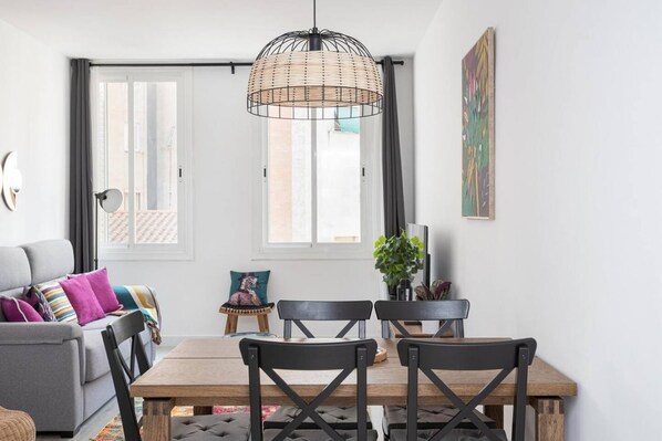 A modern living room with a grey sofa, a wooden dining table, and black chairs. There is a large window with curtains, a hanging wicker light fixture, and a potted plant.