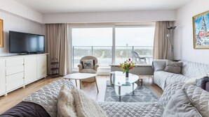 Ein modernes Wohnzimmer mit Flachbildfernseher, einer Couch, einem Sessel, einem Glastisch und Blick ins Freie.