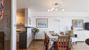 Eine moderne Küche mit weißen Schränken, einem Essbereich mit Tisch für vier Personen und einem Wohnbereich mit Sofa und Fernseher.