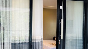 Een hotelkamer met een schuifglasdeur die leidt naar een balkon, een bed met een beige hoofdeinde, en een tafeltje met een lamp.