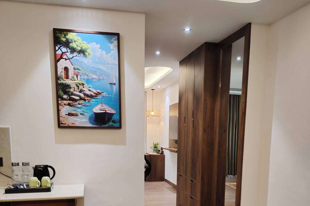 A modern hotel room with a wooden floor, a desk with a chair, a painting of a coastal scene, and a wardrobe.