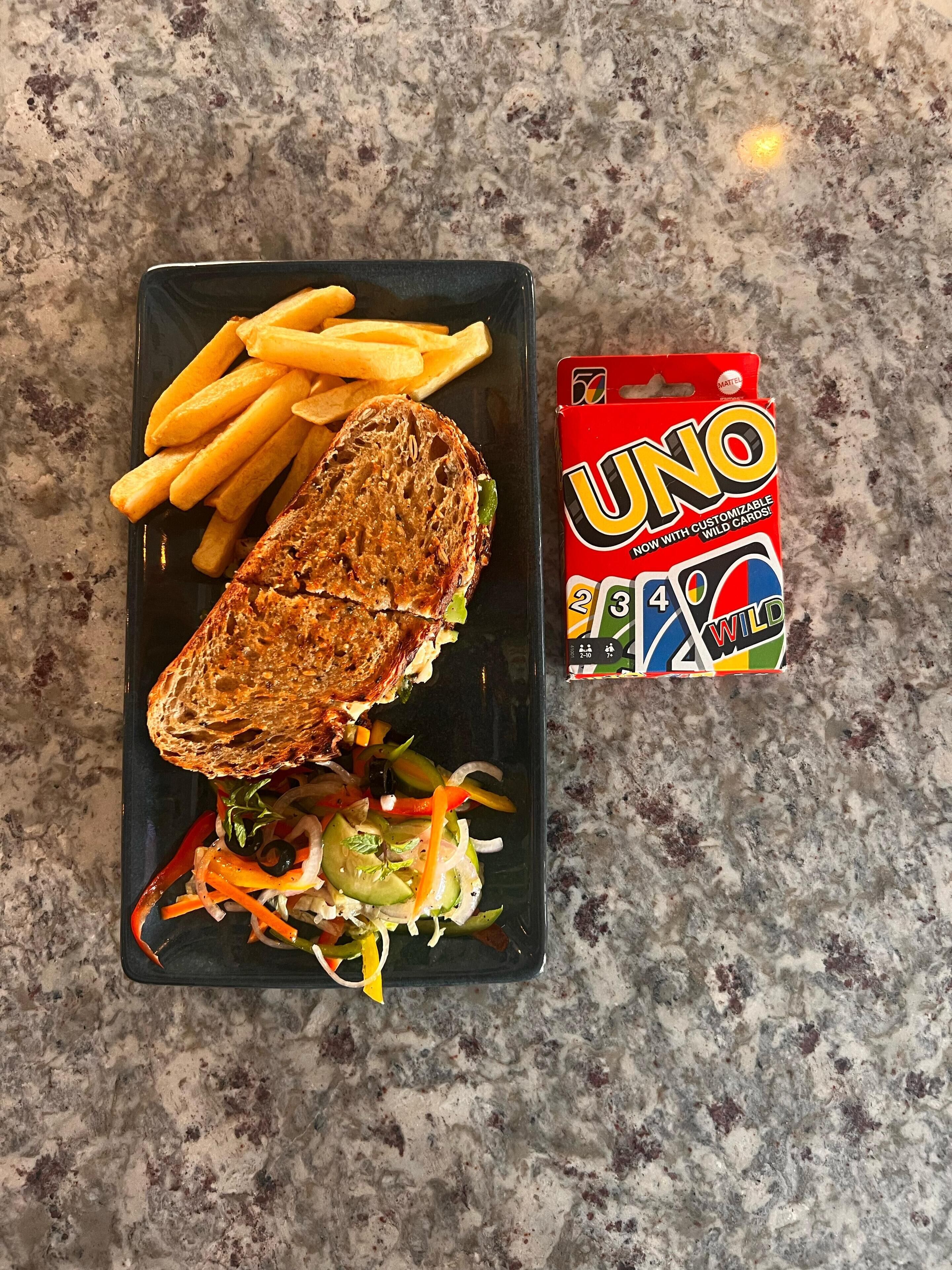 Annoksessa on grillattu voileipä, ranskalaisia perunoita ja pieni salaatti, ja se on tarjoiltu mustalla lautasella. Lautasen vieressä on UNO Wild Cards -korttipakkaus.