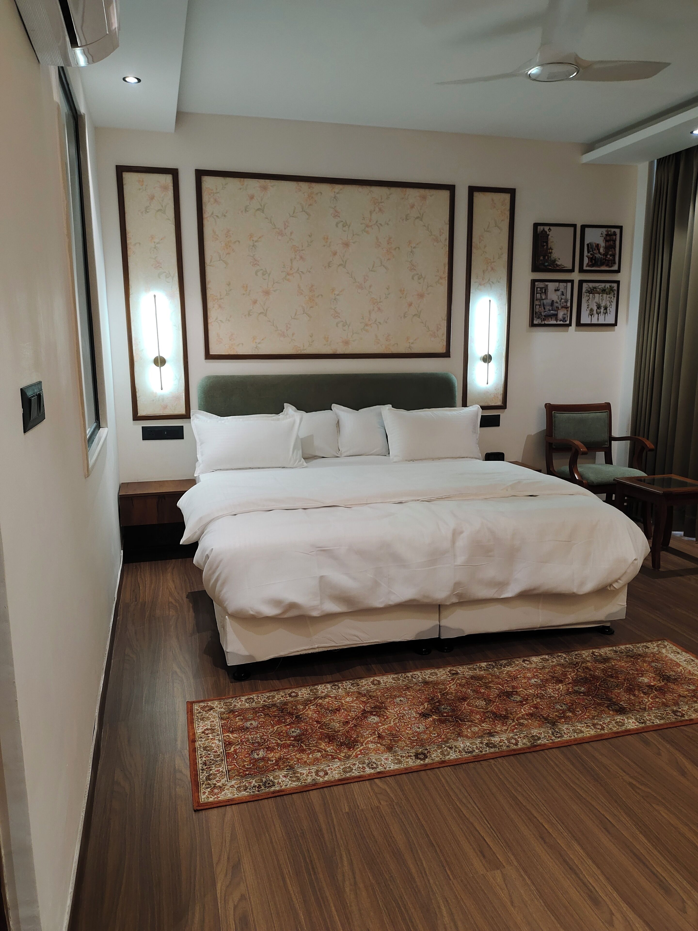 A hotel room with a large bed, a wooden floor, a decorative rug, wall-mounted lights, and framed artwork.