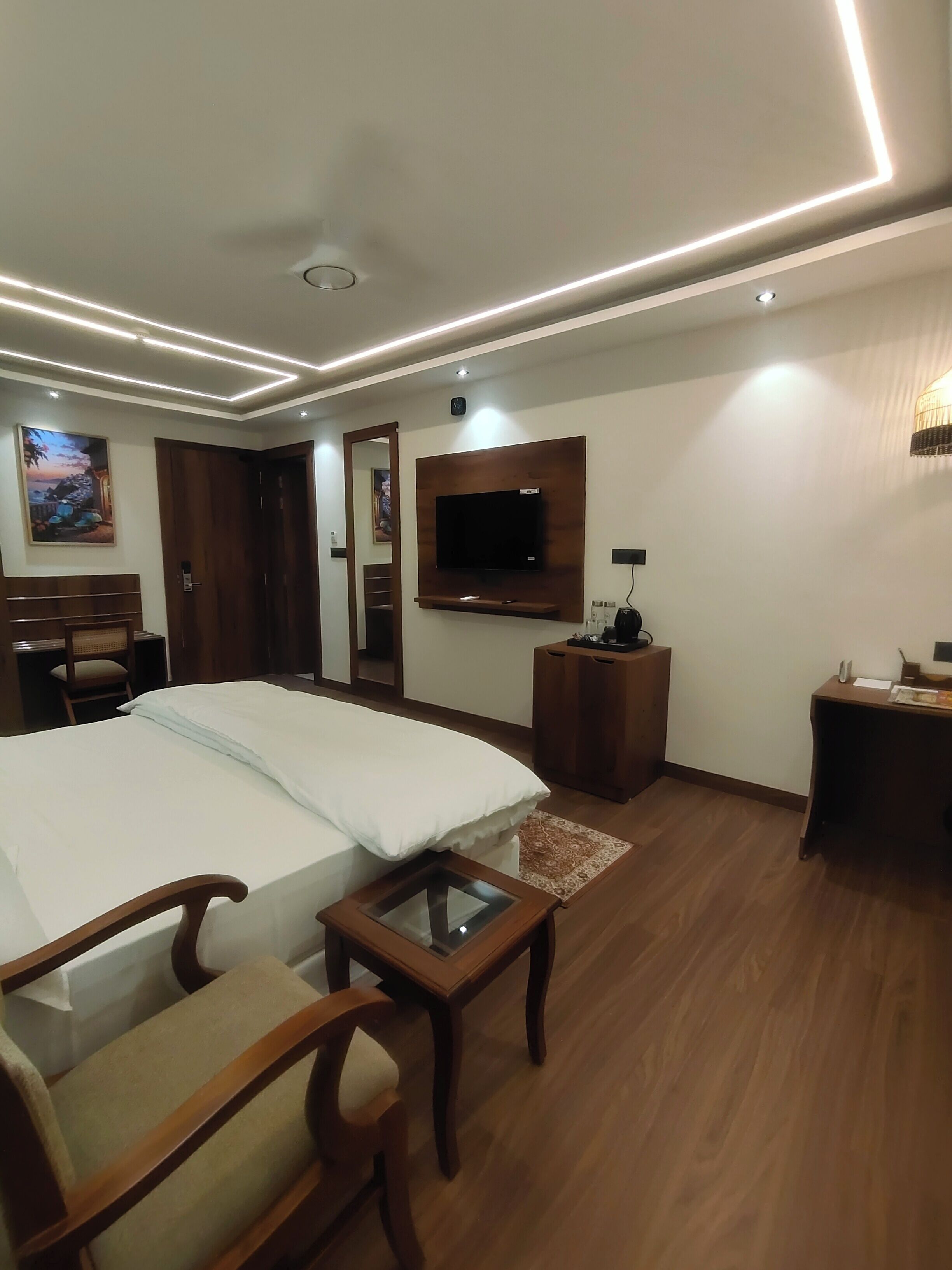 A hotel room with a bed, a desk, a chair, a TV mounted on the wall, and a wooden floor.