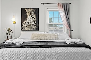 Une chambre à coucher avec une grande œuvre d’art à motifs léopard, un lit avec une literie blanche et noire, un oreiller moelleux, une table de chevet avec une lampe et une fenêtre avec des rideaux.