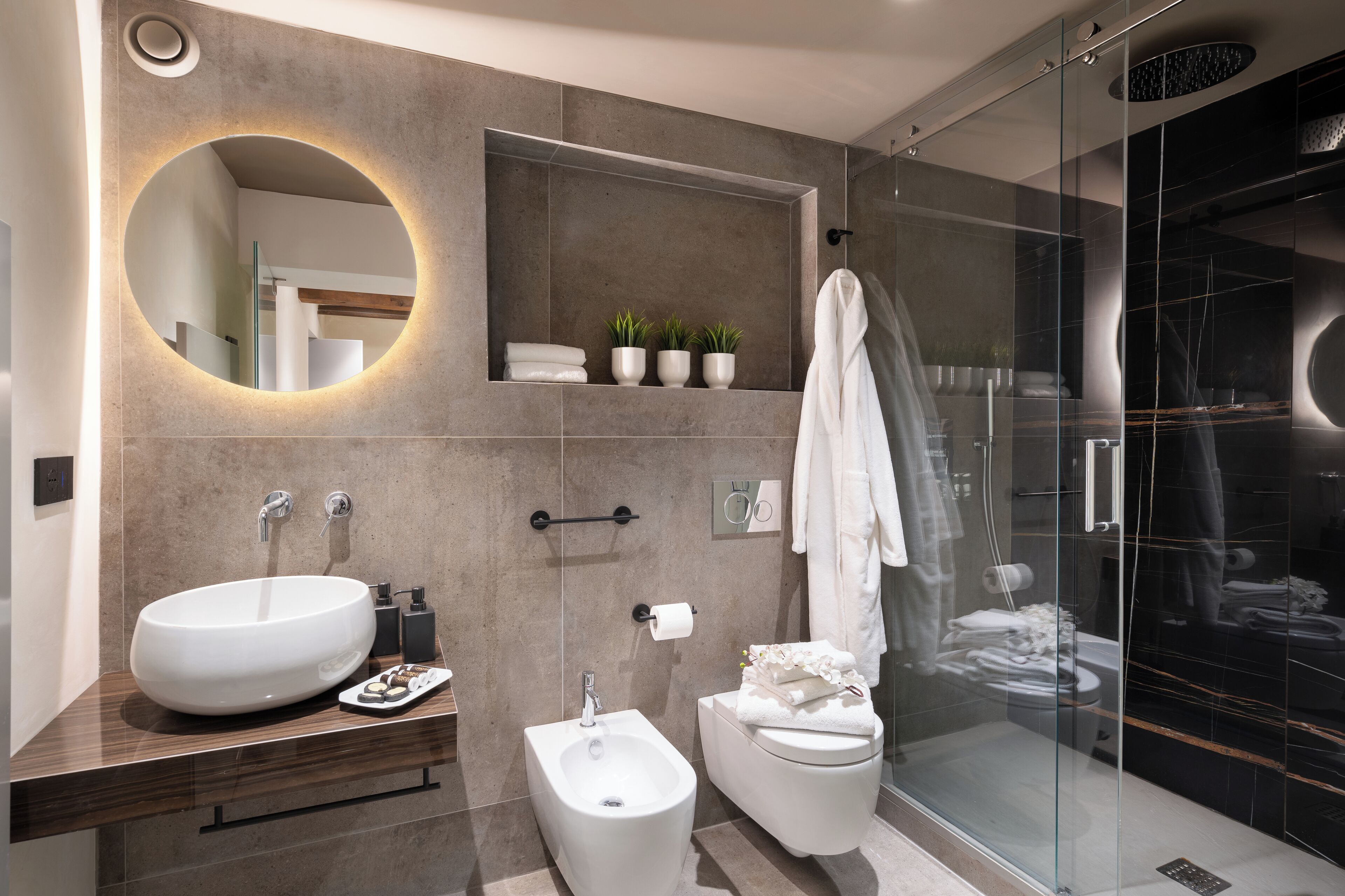 A modern bathroom with a round mirror, a large sink, a bidet, and a glass-enclosed shower.