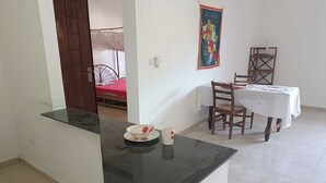 Una cocina con cubierta negra, una mesa de comedor con sillas y una cama con colcha roja al fondo.