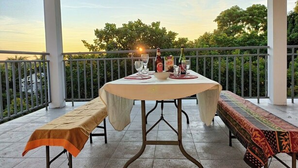 Un espacio cubierto para comer al aire libre con una mesa lista para dos, un banco y vistas a árboles y al atardecer.