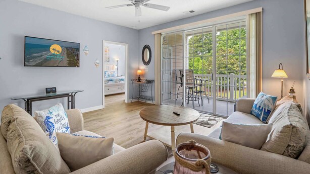 A spacious living room with a ceiling fan, a large window with a view of greenery, a dining area with a table and chairs, and a cozy seating area with a couch and pillows. - River Crossing Unit 324 (North Myrtle Beach)