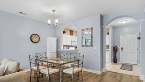 A dining area with a glass table and chairs, a chandelier, a clock, and a door leading to another room. - River Crossing Unit 324 (North Myrtle Beach)