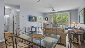A dining area with a glass table and chairs, a sofa, and a ceiling fan. - River Crossing Unit 324 (North Myrtle Beach)