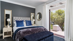 Une chambre à coucher avec une tête de lit capitonnée, deux tables de chevet, un grand lit recouvert d’une couvre-lit bleu foncé et deux miroirs muraux.