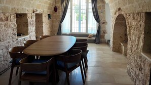 A stone-walled room with a large table and chairs, a window with curtains, and a stone bench.