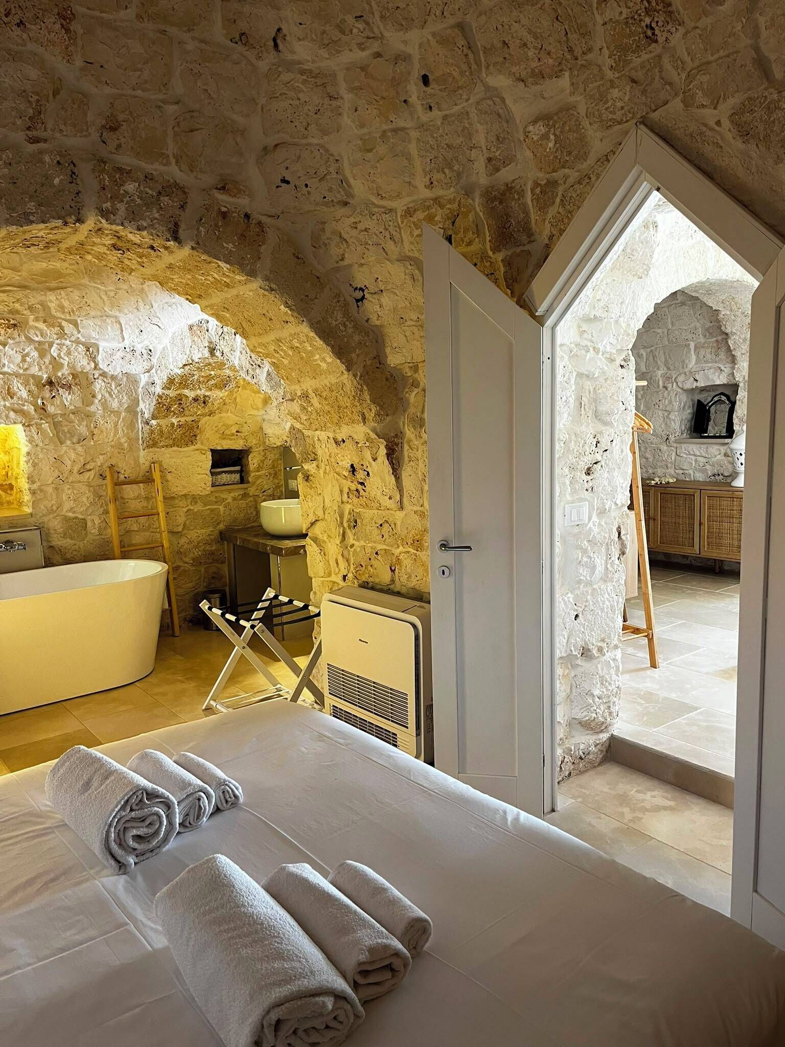 Une chambre avec un mur en pierre, une baignoire, un lit avec des serviettes et une vue sur un espace extérieur.