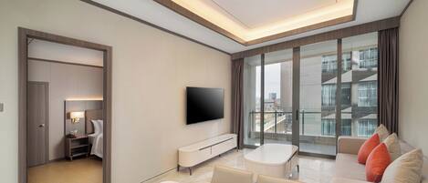 A modern hotel room with a large window, a flat-screen TV, a sofa, a dining table, and a walk-in closet.