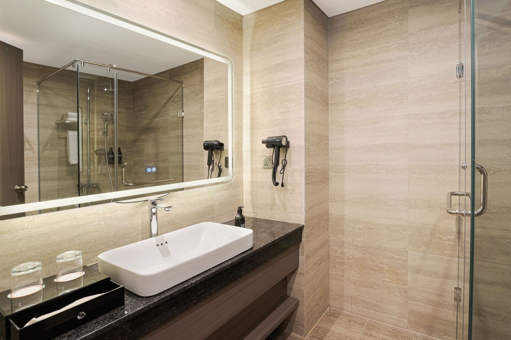 A modern bathroom with a large mirror, a white sink, a glass shower enclosure, and a hairdryer mounted on the wall.