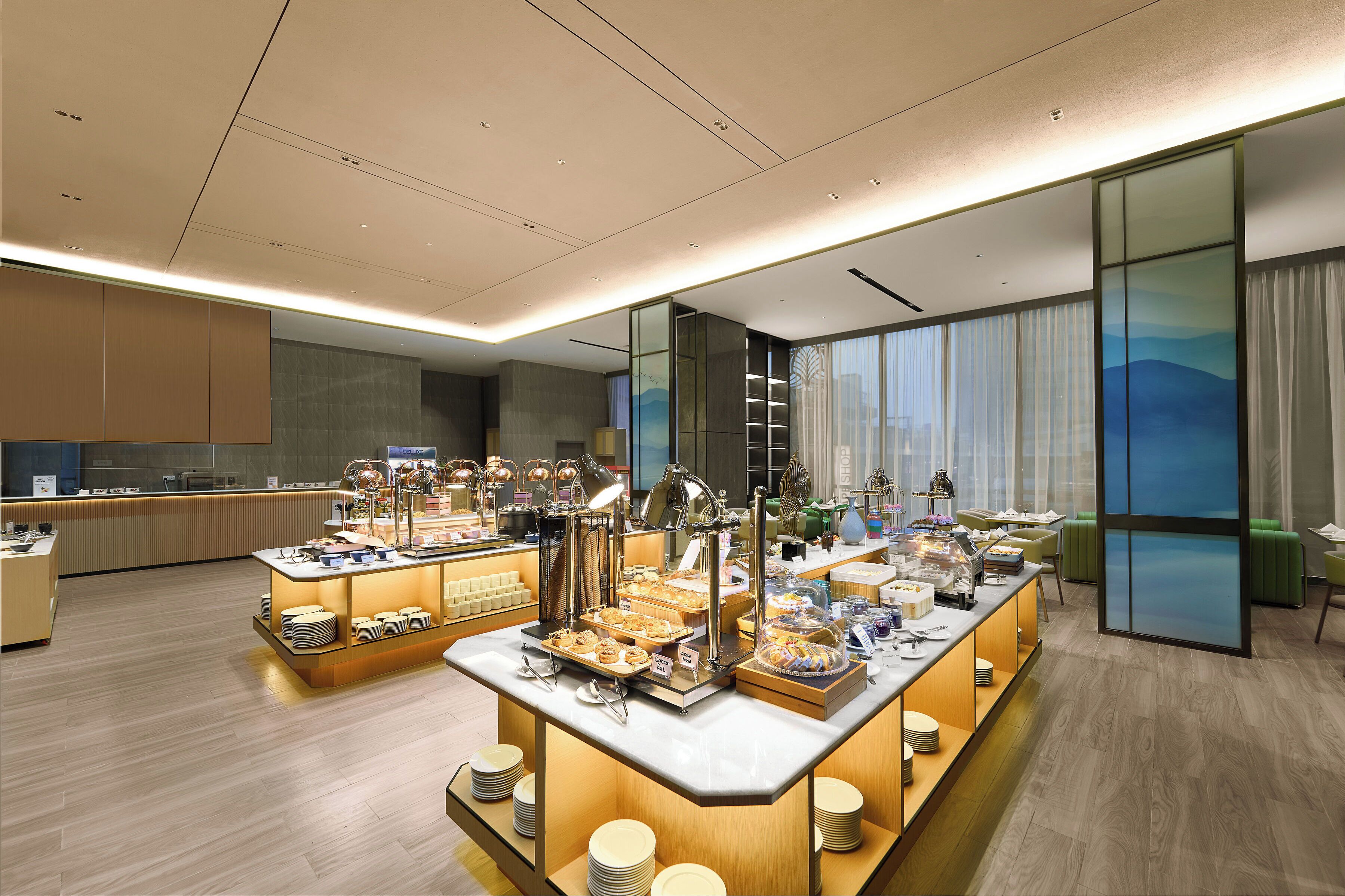A modern hotel breakfast area with multiple serving stations, a large window with a view, and a clean, contemporary design.