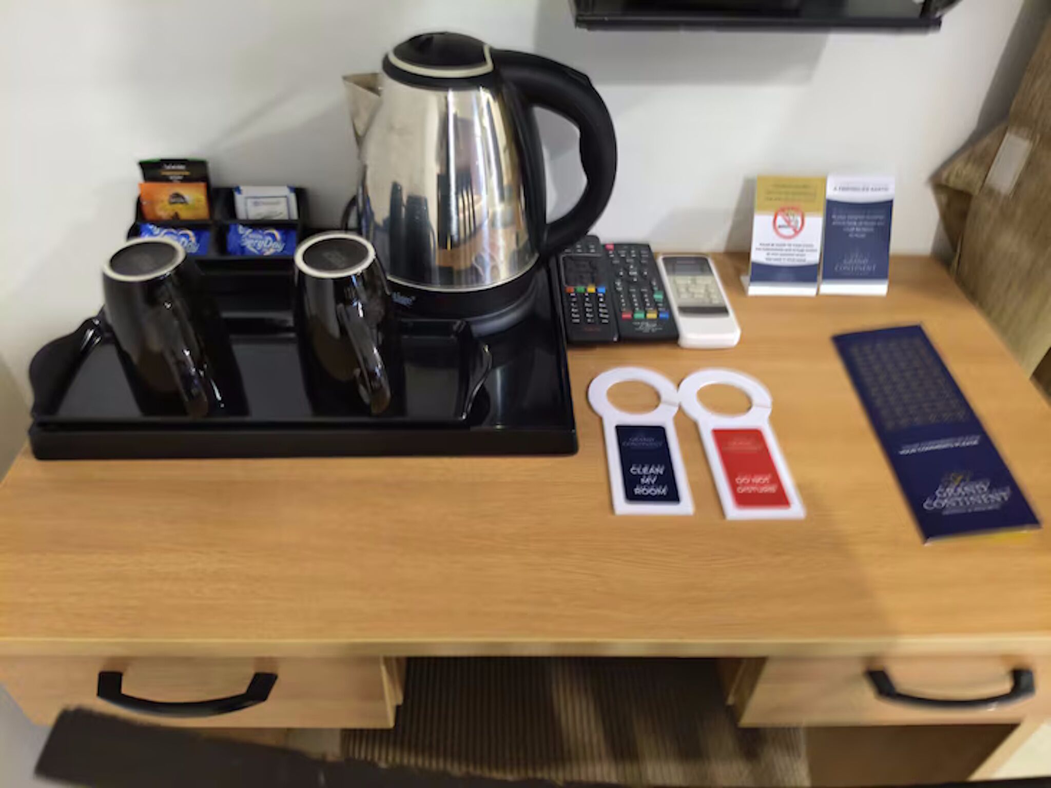 Habitación de hotel con estación de café, hervidor de agua, control remoto, teléfono, cartel de 'servicio de limpieza en la habitación' y cartel de 'no fumar'.