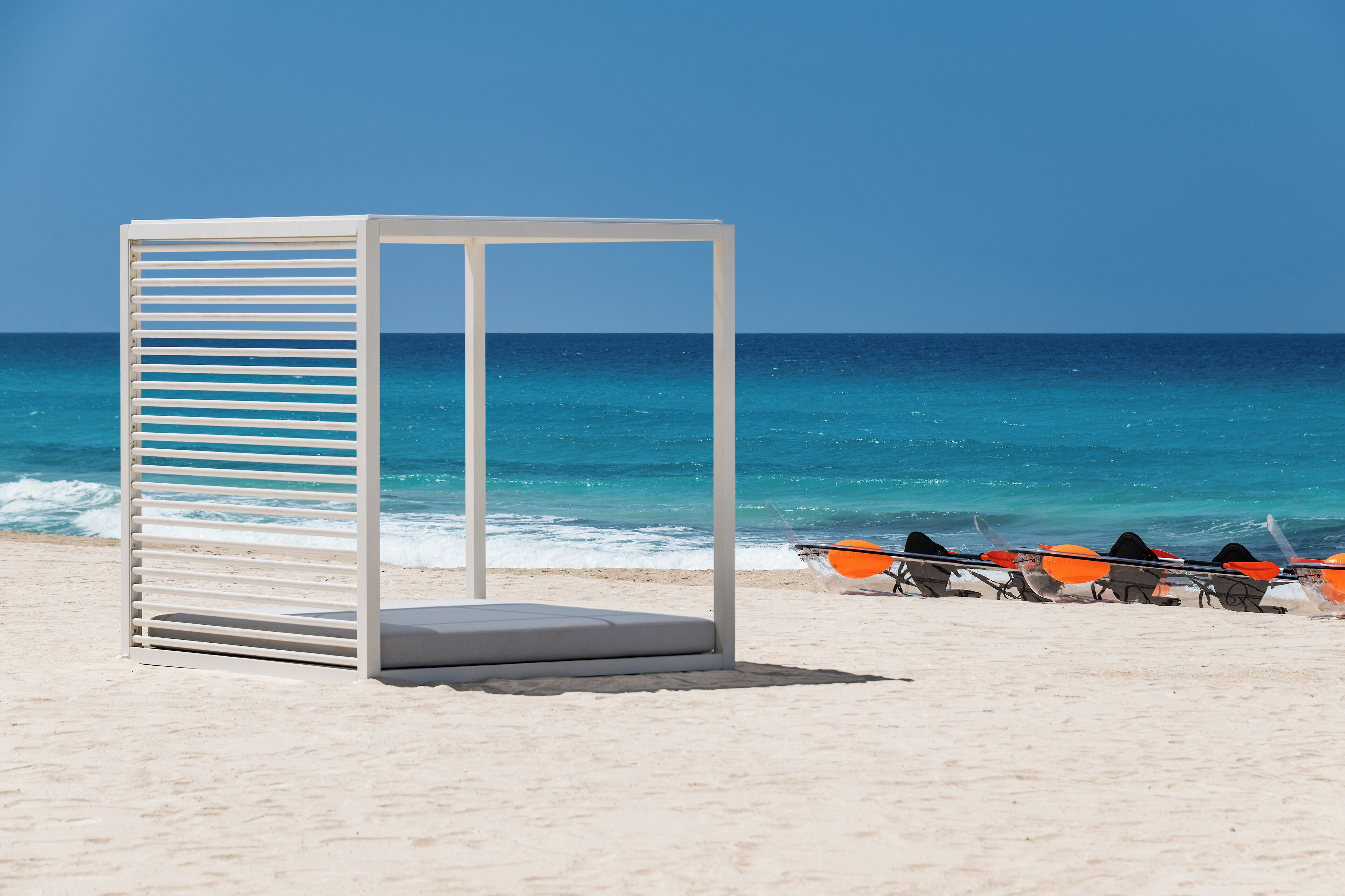A beach with a white structure, possibly a lounge or seating area, and a row of orange paddleboards.