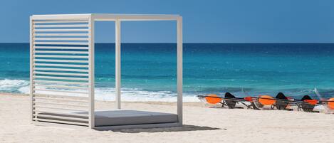A beach with a white structure, possibly a lounge or seating area, and a row of orange paddleboards.