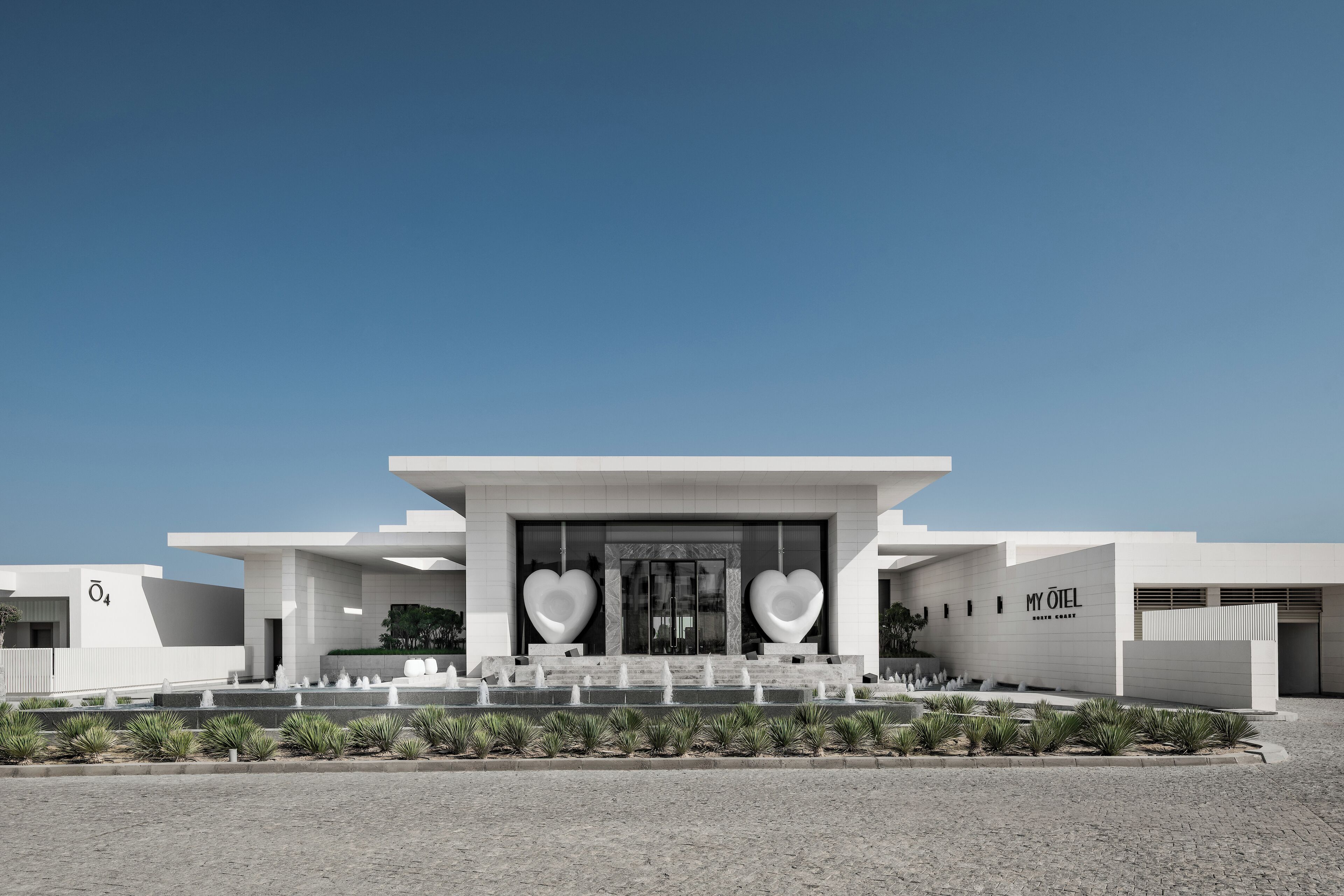 A modern building with a large heart-shaped sculpture in front, a clear sky, and a paved area with plants.