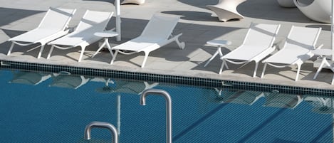 A pool area with white lounge chairs and umbrellas, a clear blue pool, and a tiled deck.
