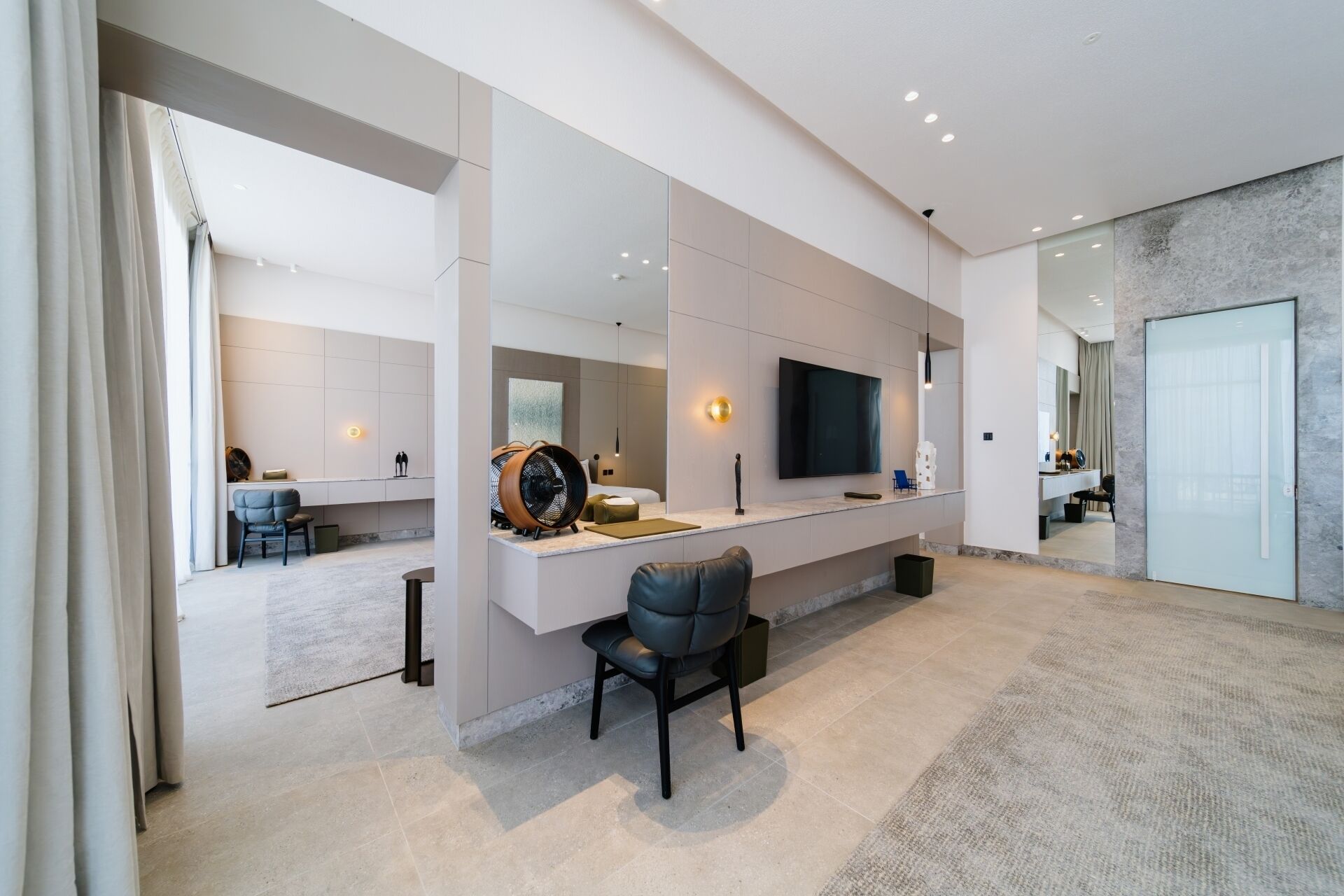 Une chambre d’hôtel moderne dotée d’une grande télévision à écran plat, d’un coin salon avec un canapé et des fauteuils, d’un grand miroir et d’un sol recouvert de carreaux de marbre.