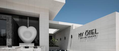 A modern hotel entrance with a large heart-shaped sculpture, a pool area, and the name 'MY OTEI NORTH COAST' on the wall.