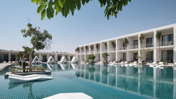 A swimming pool with white lounge chairs and palm trees, surrounded by a modern building with balconies. - My Ōtel North Coast (El Dabaa)
