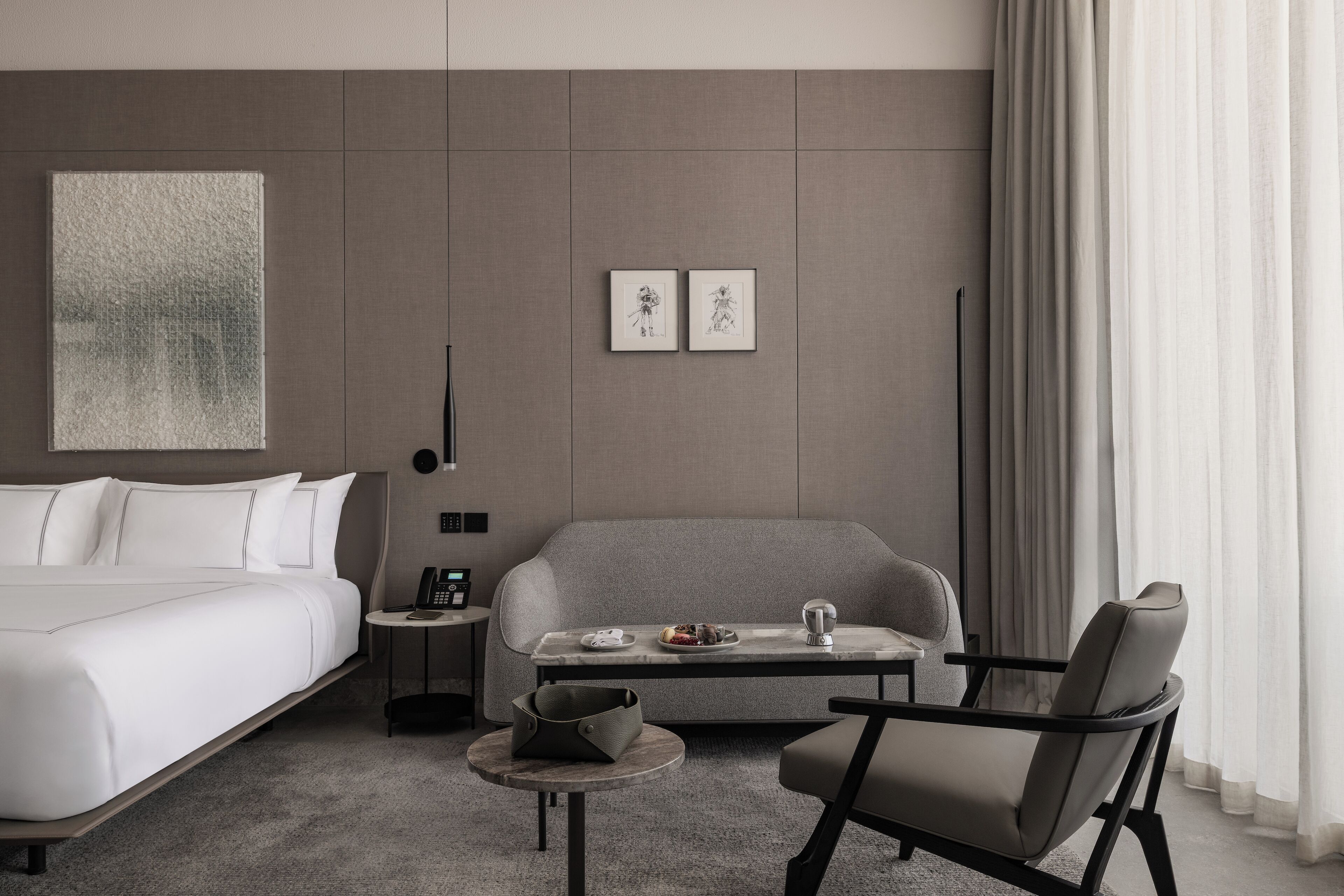 Une chambre d’hôtel moderne avec un lit, un canapé, une petite table avec un téléphone et un sac, et deux tableaux encadrés au mur.
