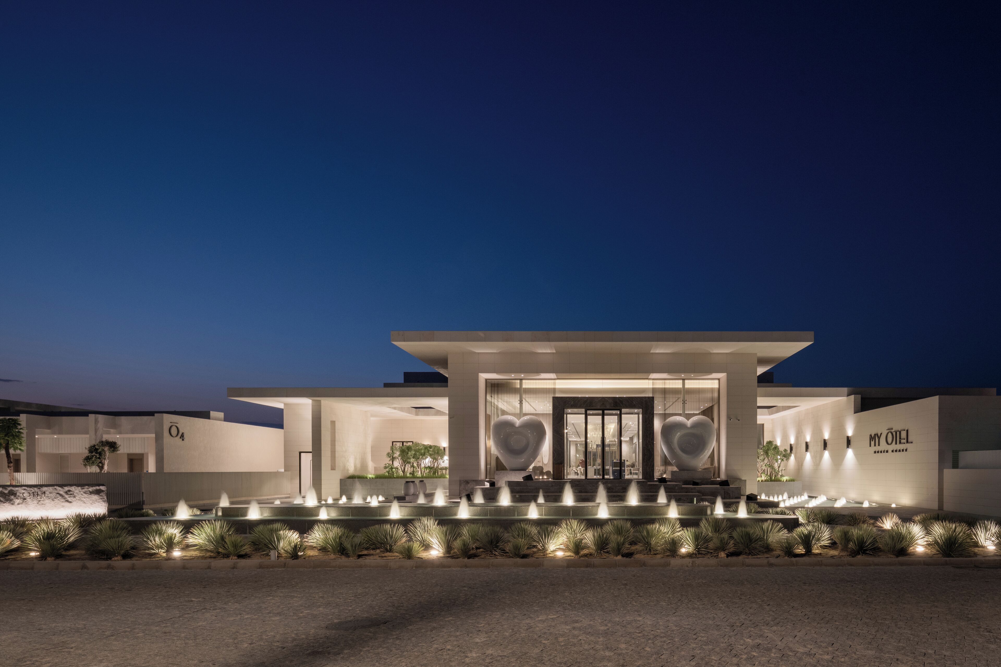 A modern hotel entrance with a glass facade, a heart-shaped sculpture, and a well-lit fountain area.