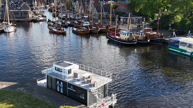 Un port de plaisance où sont amarrés différents types de bateaux, une petite structure flottante dotée d’une terrasse et de sièges, et une ville en arrière-plan.