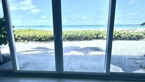 A view through large windows of a concrete patio with a beach and ocean in the background.