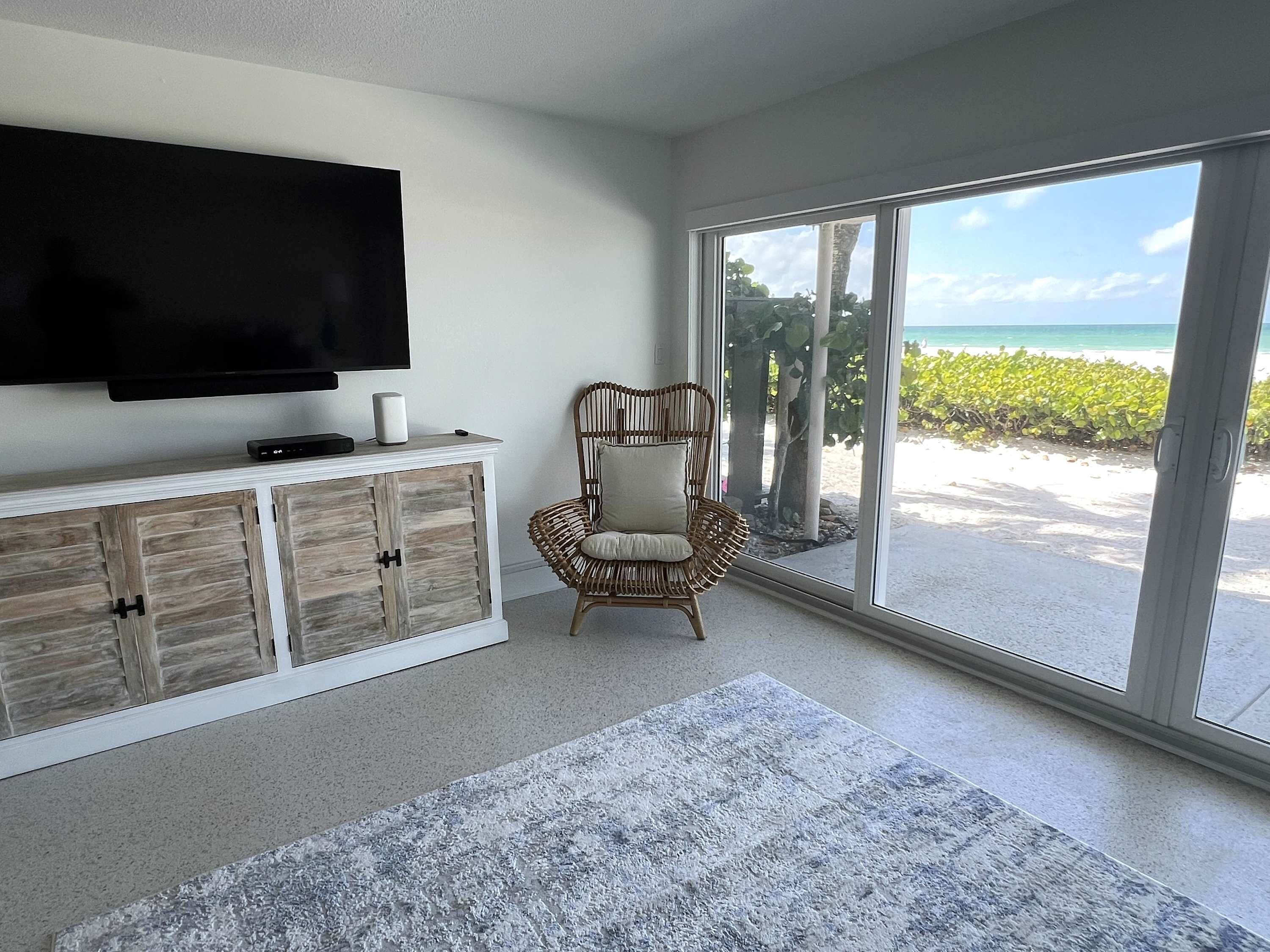 Una habitación con un televisor grande sobre un mueble de madera, un sillón de mimbre y vistas a la playa a través de puertas correderas de cristal.