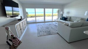 A spacious living room with a large window offering a view of the beach, a flat-screen TV mounted on the wall, and a white sofa with blue cushions.