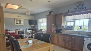 A modern kitchen with wooden cabinets, a dining area with a table and chairs, a built-in washing machine, and a large window with a view. - Tranquil Cottage Retreat in Kells, Leitrim (Mullagh)