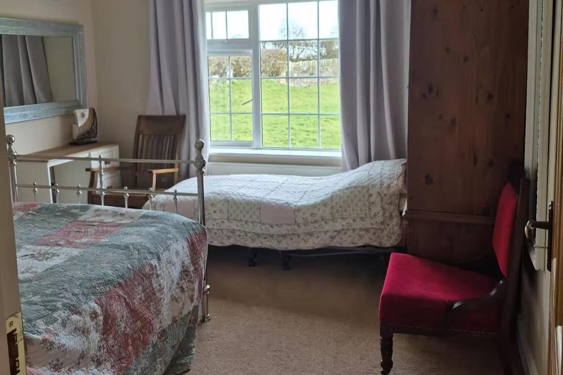 A bedroom with two beds, a chair, a window with curtains, and a mirror.