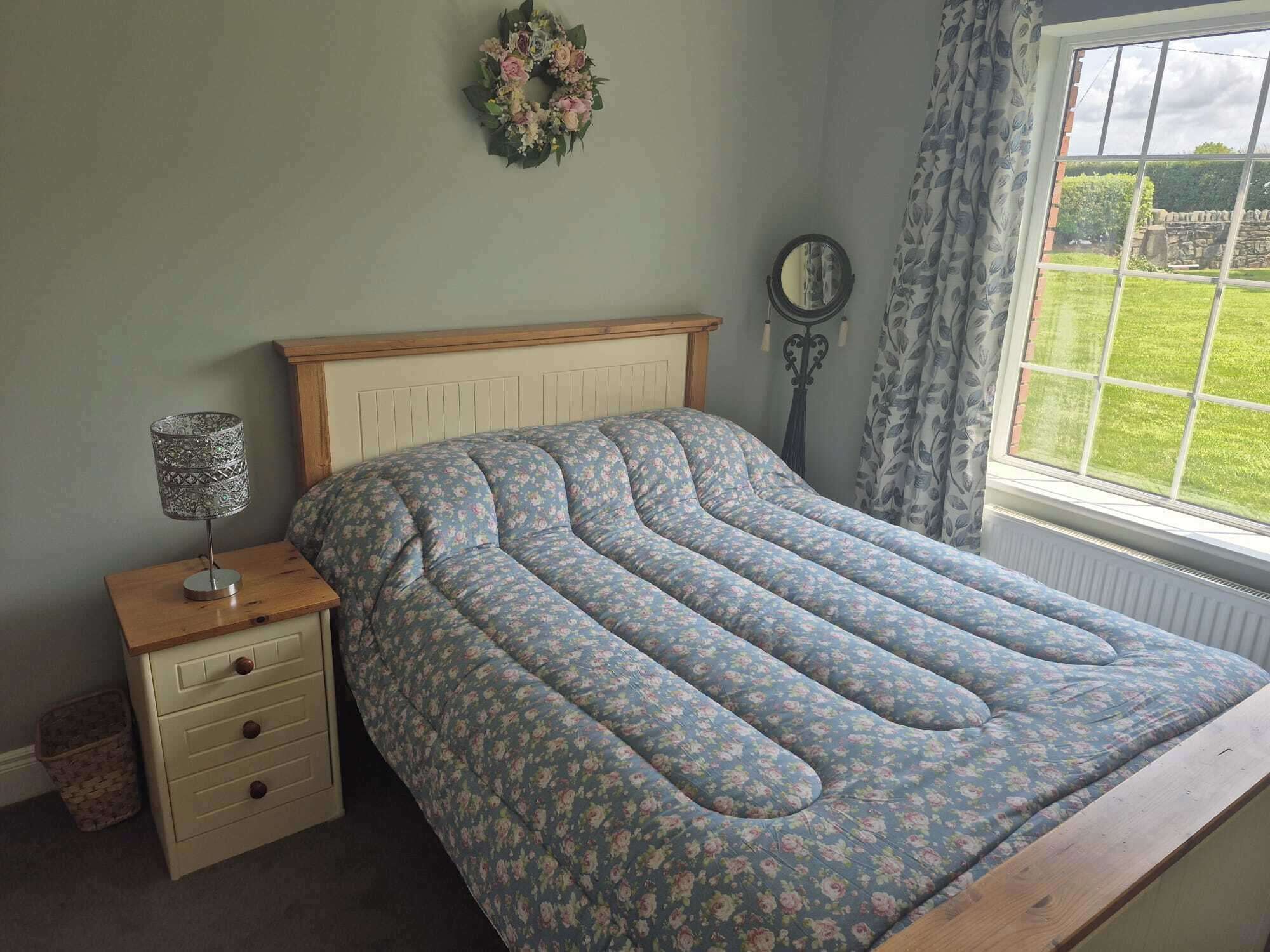 A bedroom with a floral-patterned bed, a bedside table with a lamp, a mirror, and a view of a garden through a window.