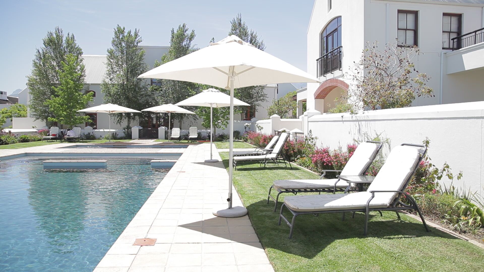 A swimming pool with lounge chairs and umbrellas, surrounded by a well-maintained lawn and a building with a white facade.