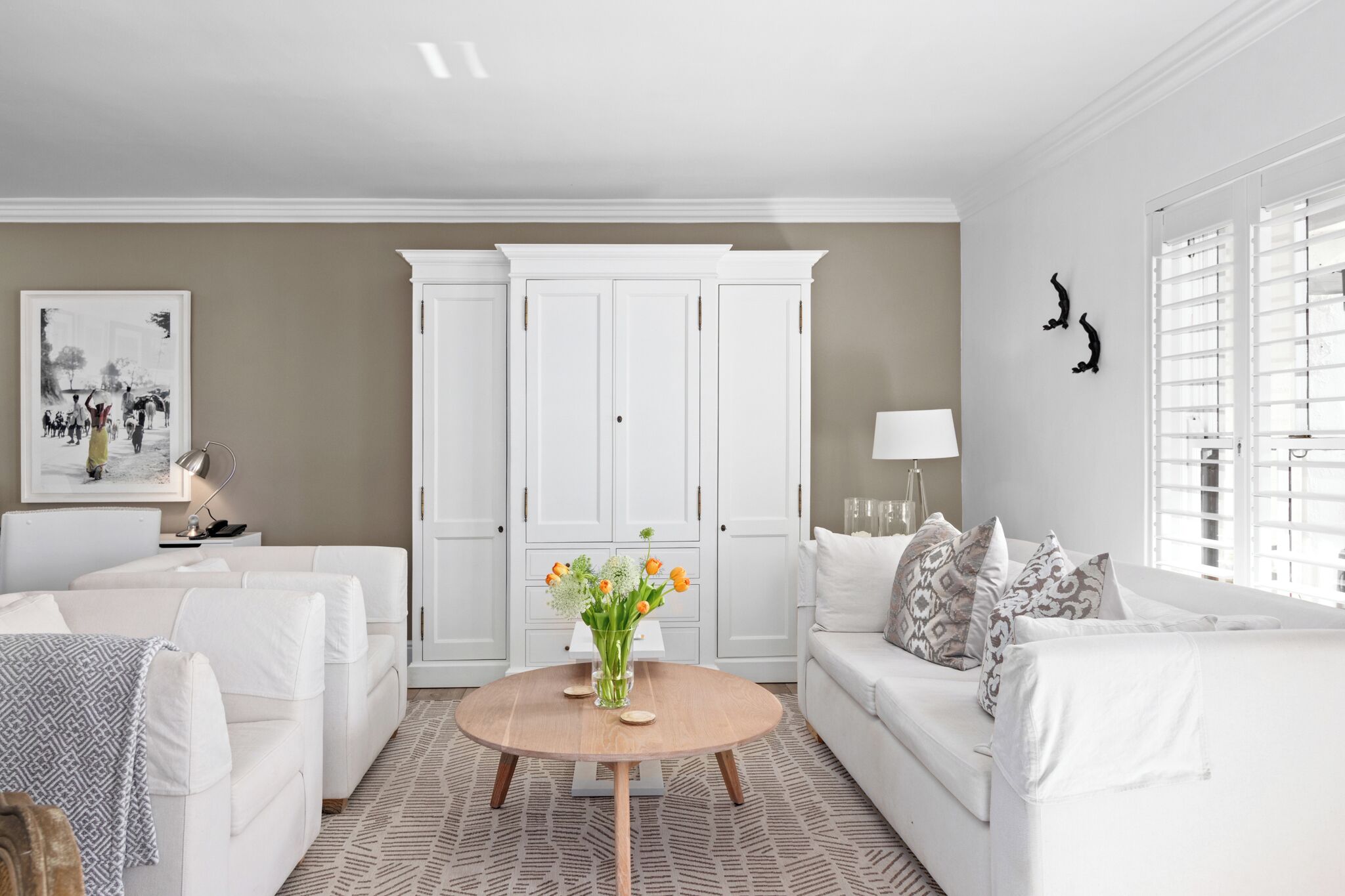 A modern living room with white furniture, a round wooden coffee table, and a beige accent wall.