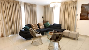 A modern living room with a black sectional sofa, two gold wire chairs, a glass coffee table, and beige curtains.