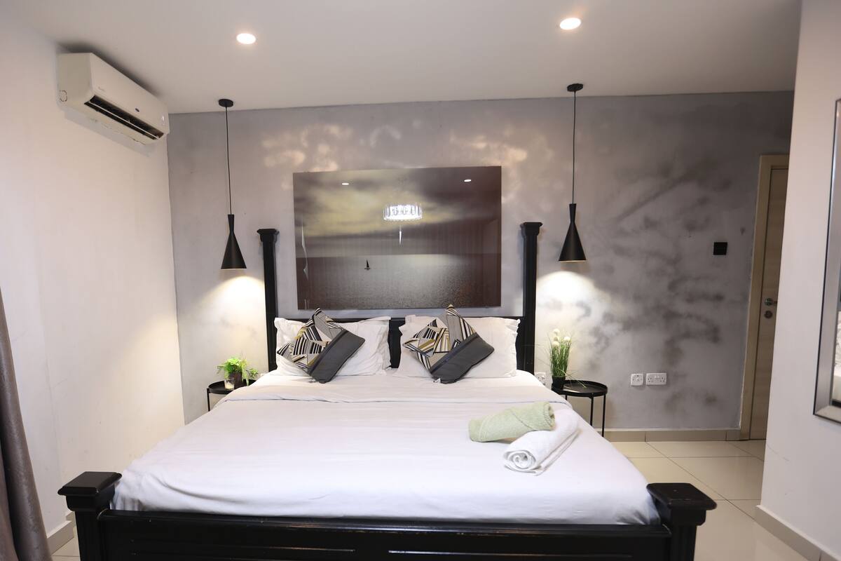 Een moderne slaapkamer met een groot bed, een flatscreen televisie aan de wand, hanglampen en een klein bijzettafeltje met een plant.