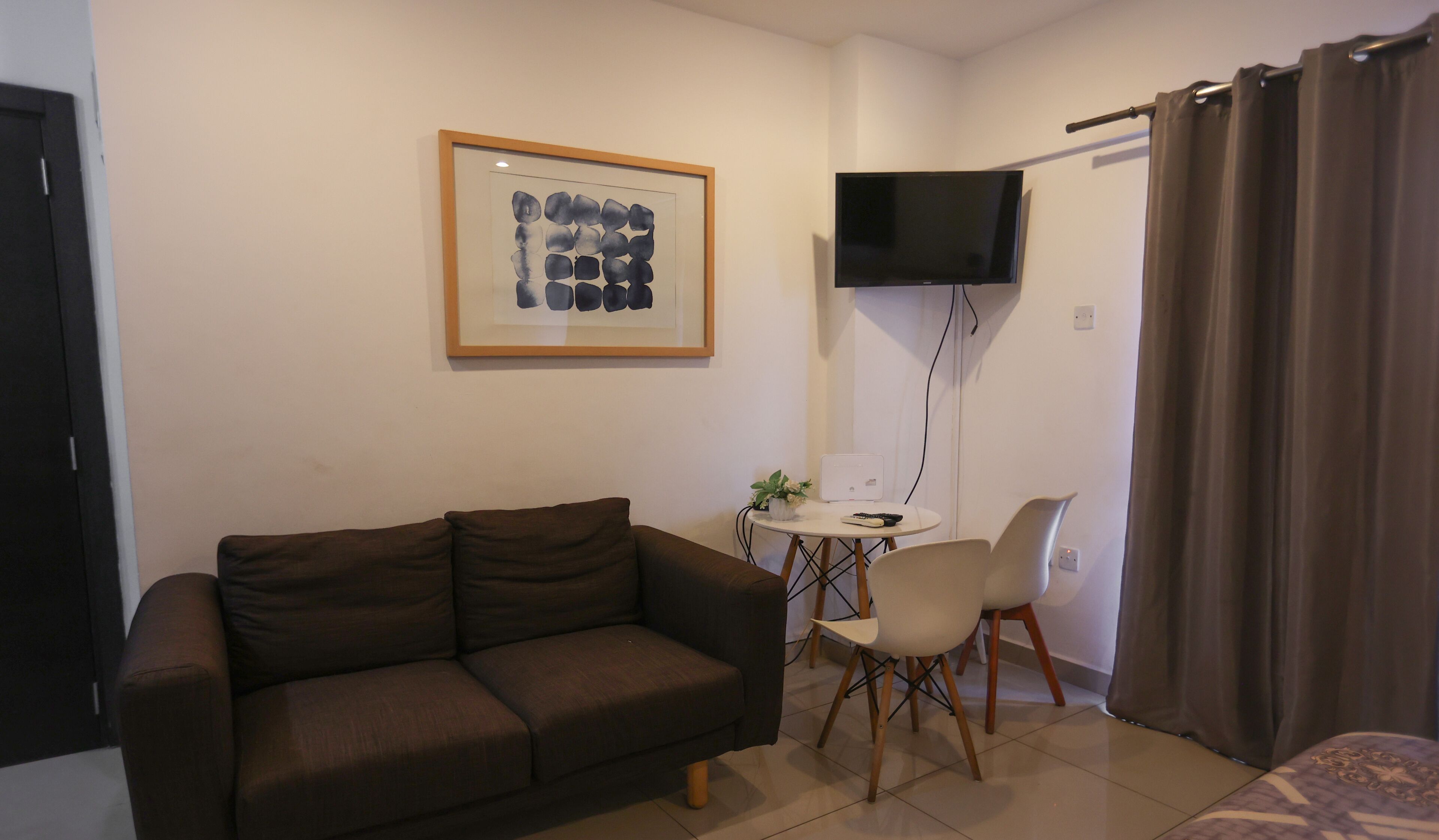 Uma sala de estar com um sofá marrom, uma mesinha com uma cadeira branca, uma obra de arte emoldurada na parede e uma TV fixada acima da mesa.