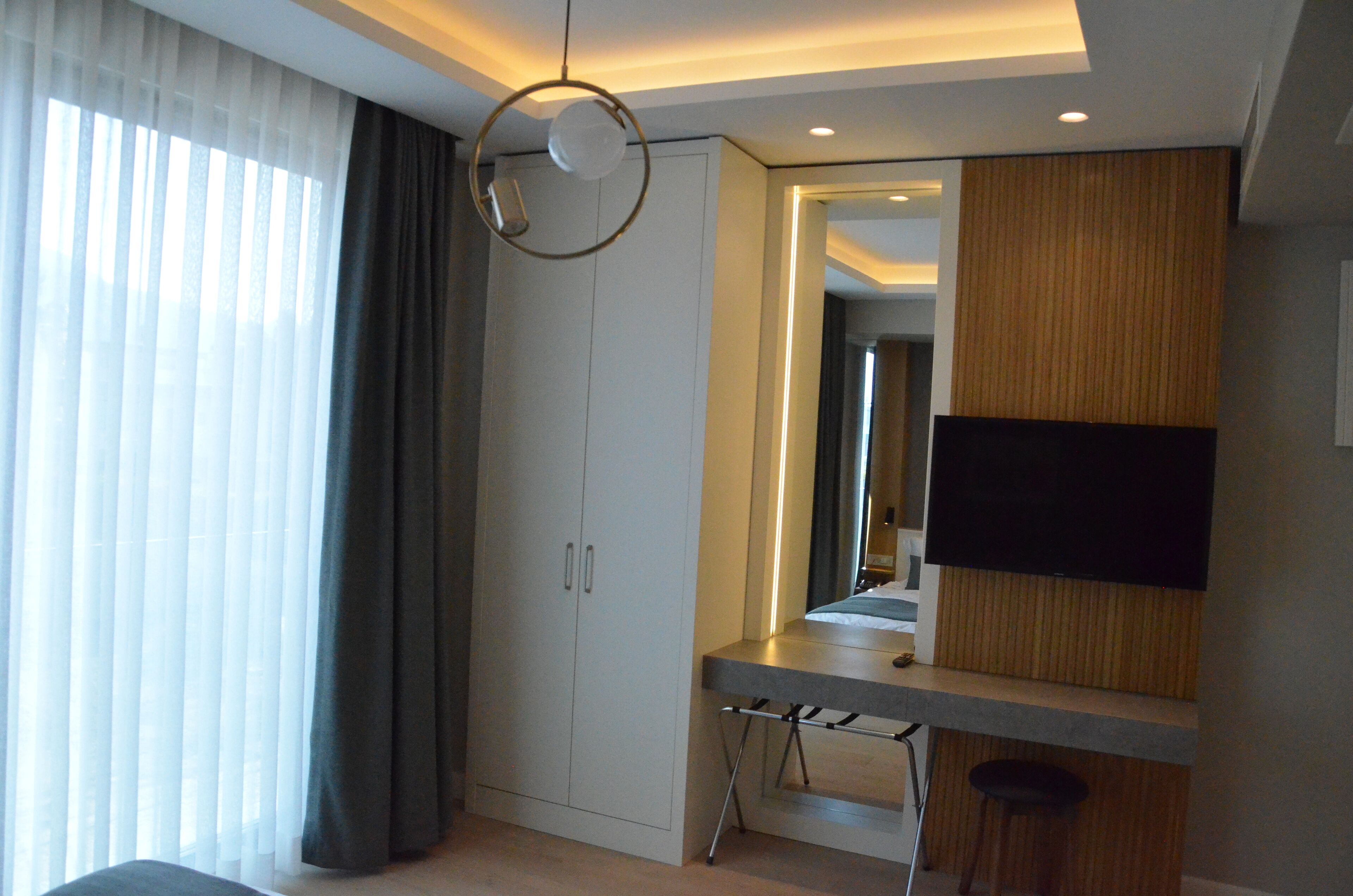 Ett modernt hotellrum med en stor garderob, ett skrivbord med en stol, en platt-TV som är monterad på väggen och ett stort fönster med tunna, genomskinliga gardiner.