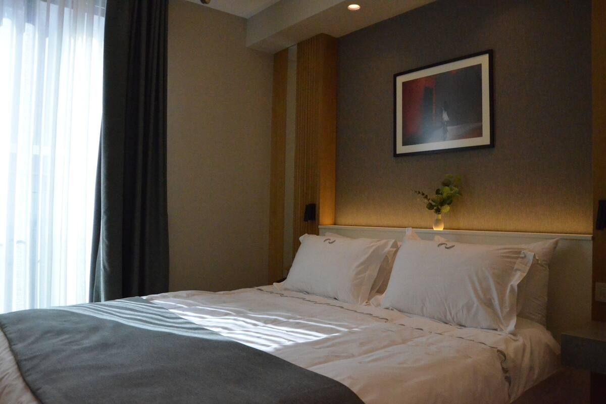 Quarto de hotel com cama, mesa de cabeceira, quadro na parede e janela com cortinas.