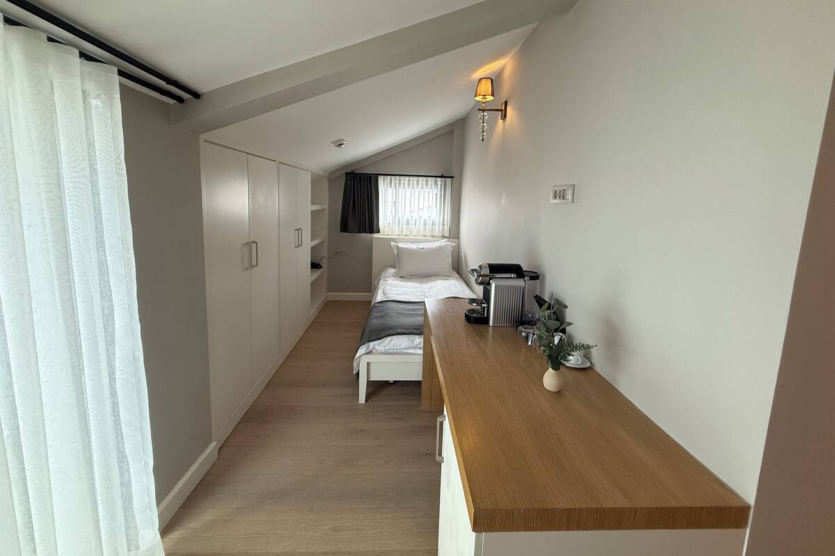 Um quarto pequeno e bem iluminado, com uma cama, uma escrivaninha de madeira, um radiador e uma luminária na parede.