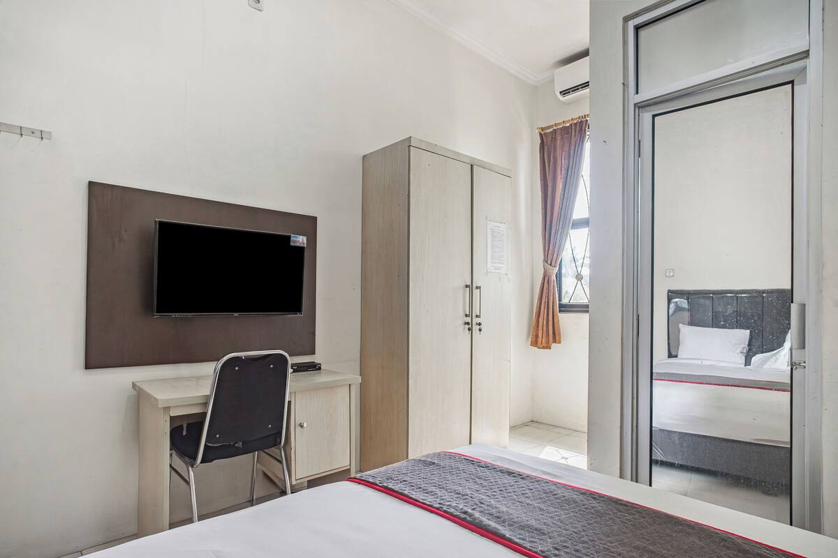 Habitación de hotel con cama, escritorio con silla, armario, televisor montado en la pared y puerta corrediza de vidrio que da a un balcón.