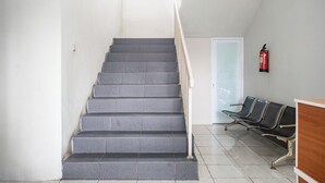 Eine Treppe mit grauen Stufen und einem weißen Geländer führt zu einer Tür. An der Wand stehen zwei schwarze Stühle mit Metallgestellen. Ein roter Feuerlöscher ist rechts an der Wand befestigt.
