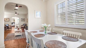 Un comedor con una mesa blanca, individuales tejidos y sillas blancas. Hay una araña sobre la mesa y una ventana con persianas a la derecha.