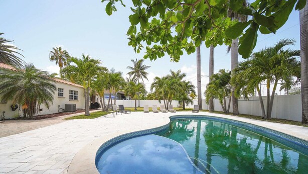 Una piscina de borde curvo, rodeada por un patio empedrado con sillas de descanso y palmeras.